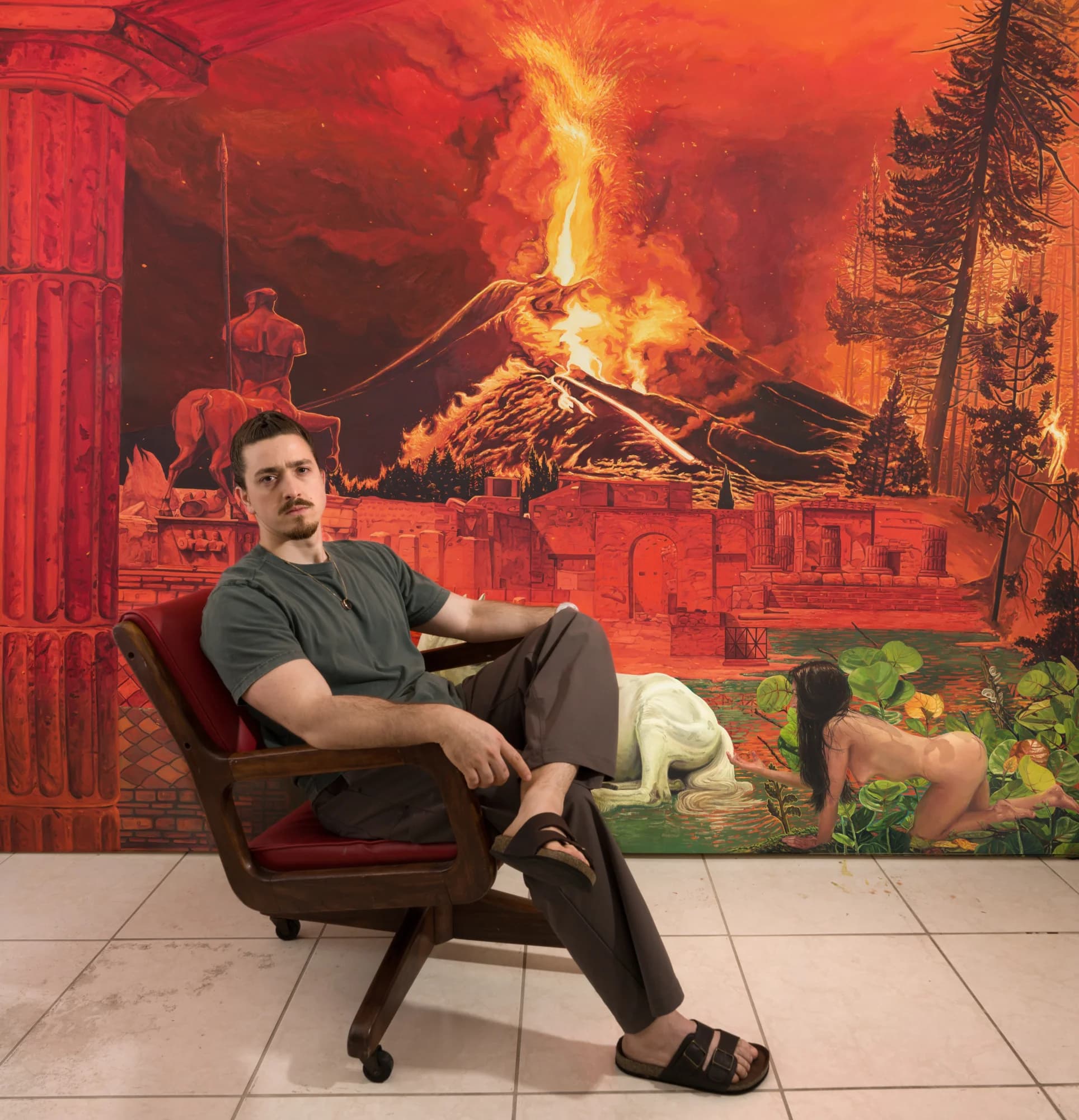 Un hombre con camiseta verde grisácea sentado en un sillón de madera sobre un piso de baldosas, frente a una gran pintura roja y naranja de un volcán en erupción sobre ruinas clásicas, con un caballo blanco y una mujer a gatas abajo a la derecha.